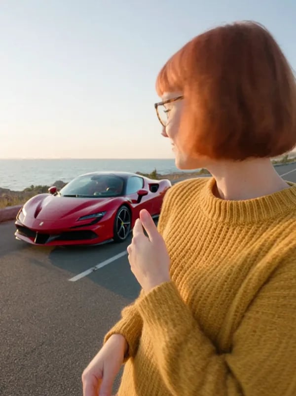 A woman with a red bob and glasses in a yellow sweater on a sunset coast road, a red Ferrari SF90 parked behind her, from the Kling test run