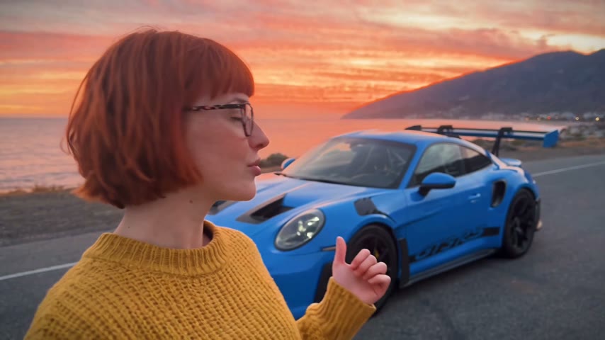 A woman in a mustard sweater talks to the camera on a sunset coast road with a blue Porsche 911 GT3 RS parked behind her